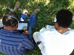 Two people sit on the grass, wearing headphones and looking at a tablet screen.