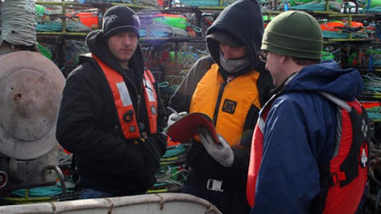 Three people in safety vests and knit hats stand together while one checks a notebook.