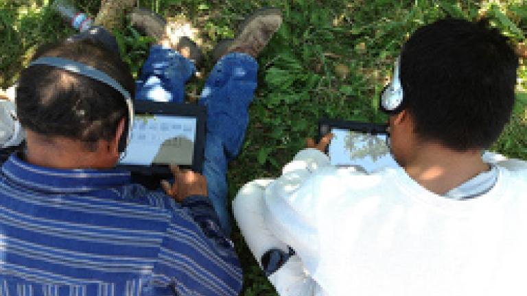 Two people sit on the grass, wearing headphones and looking at a tablet screen.