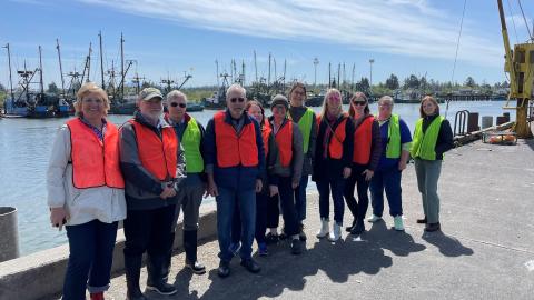 A group of people pose for a photo near a fishing harbor. They each wear orange safety vests.
